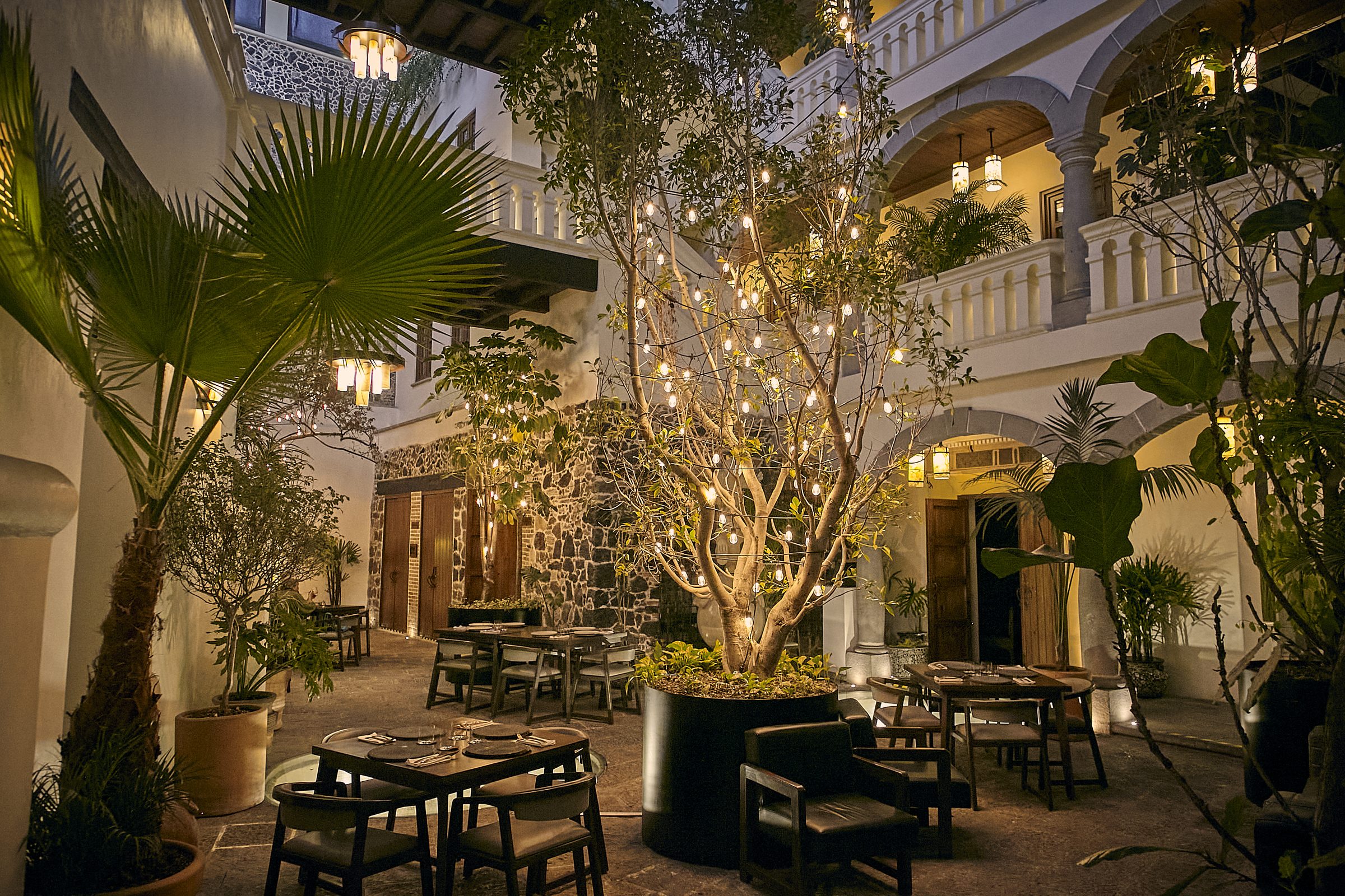 Patio central de la Casona de los Sapos de noche, con un árbol iluminado, mesas del restaurante y arcos de cantera.