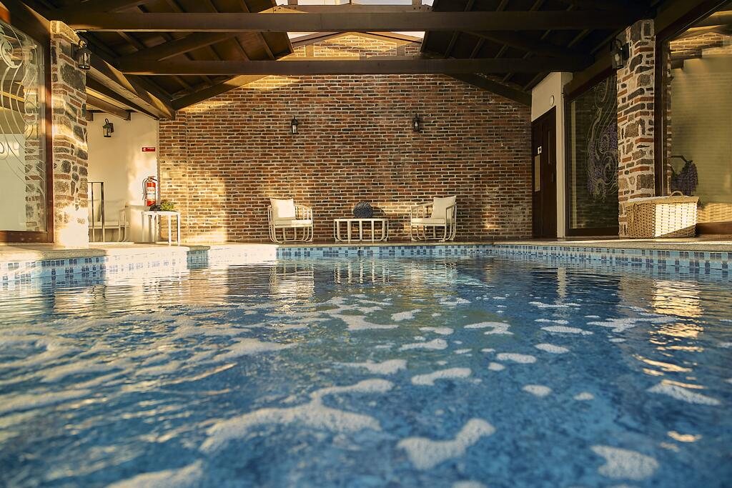 Alberca techada con muro de ladrillo, columnas de piedra y sillones junto al agua.