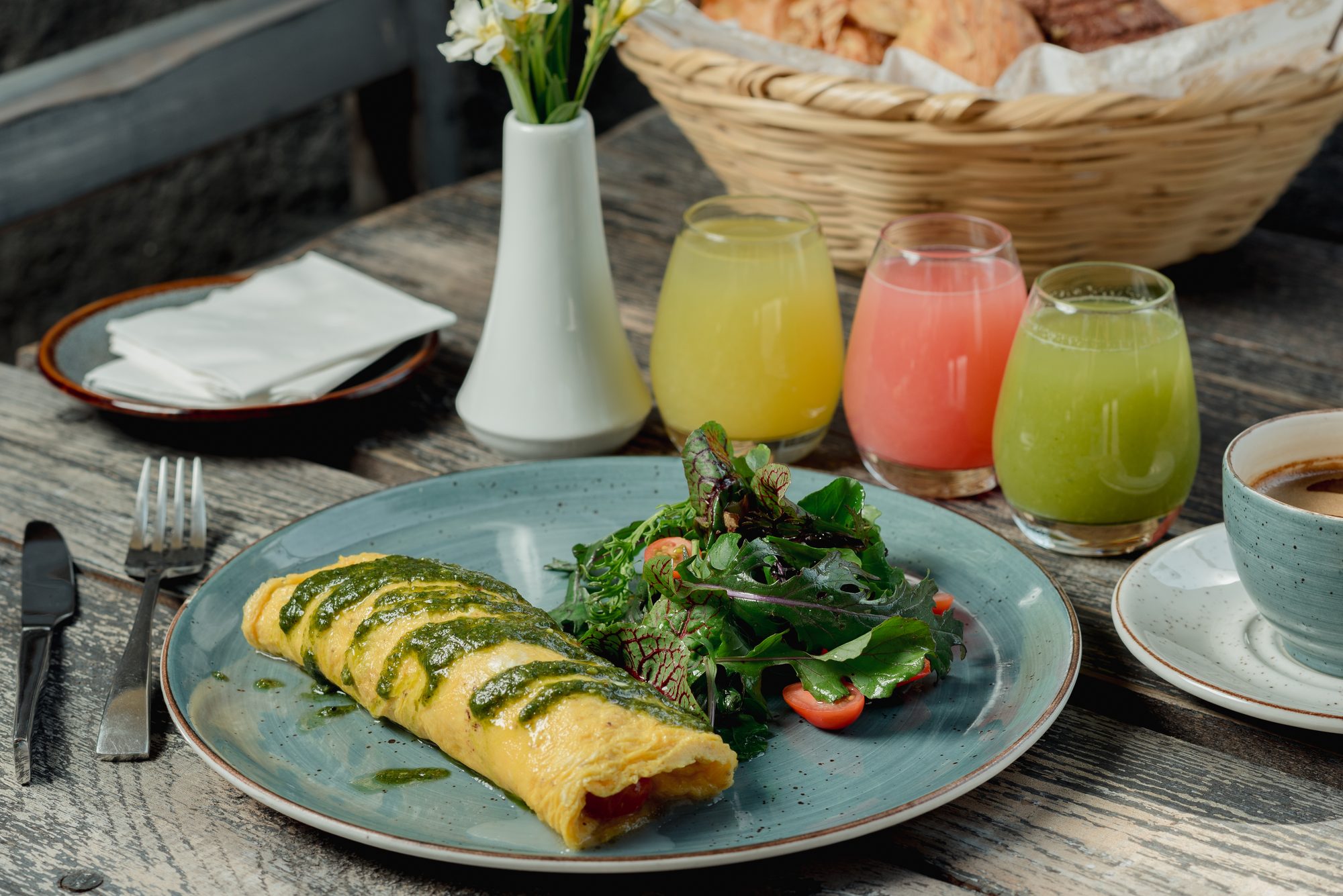 Omelette con salsa verde, ensalada, jugos y café sobre una mesa de madera, con una canasta de pan al fondo.